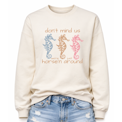 Bold summer coastal graphic on a classic off white crewneck sweatshirt. The design features three ornamental seahorses in soft coastal colors with the wording don't mind us were just horse'n around. 
