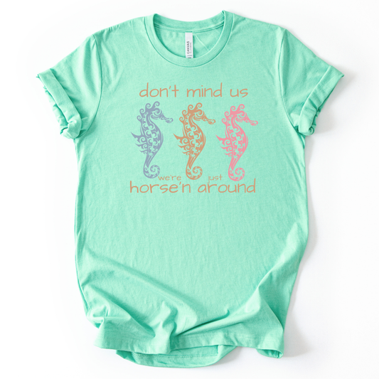 Coastal sea life graphic on a soft unisex heather mint graphic tee. The design features three decorative seahorse illustrations arranged across the center of the artwork in soft coastal colors including blue, coral, and peach. Each seahorse contains detailed swirl and ocean pattern elements that give the sea life illustration a textured coastal aesthetic. 