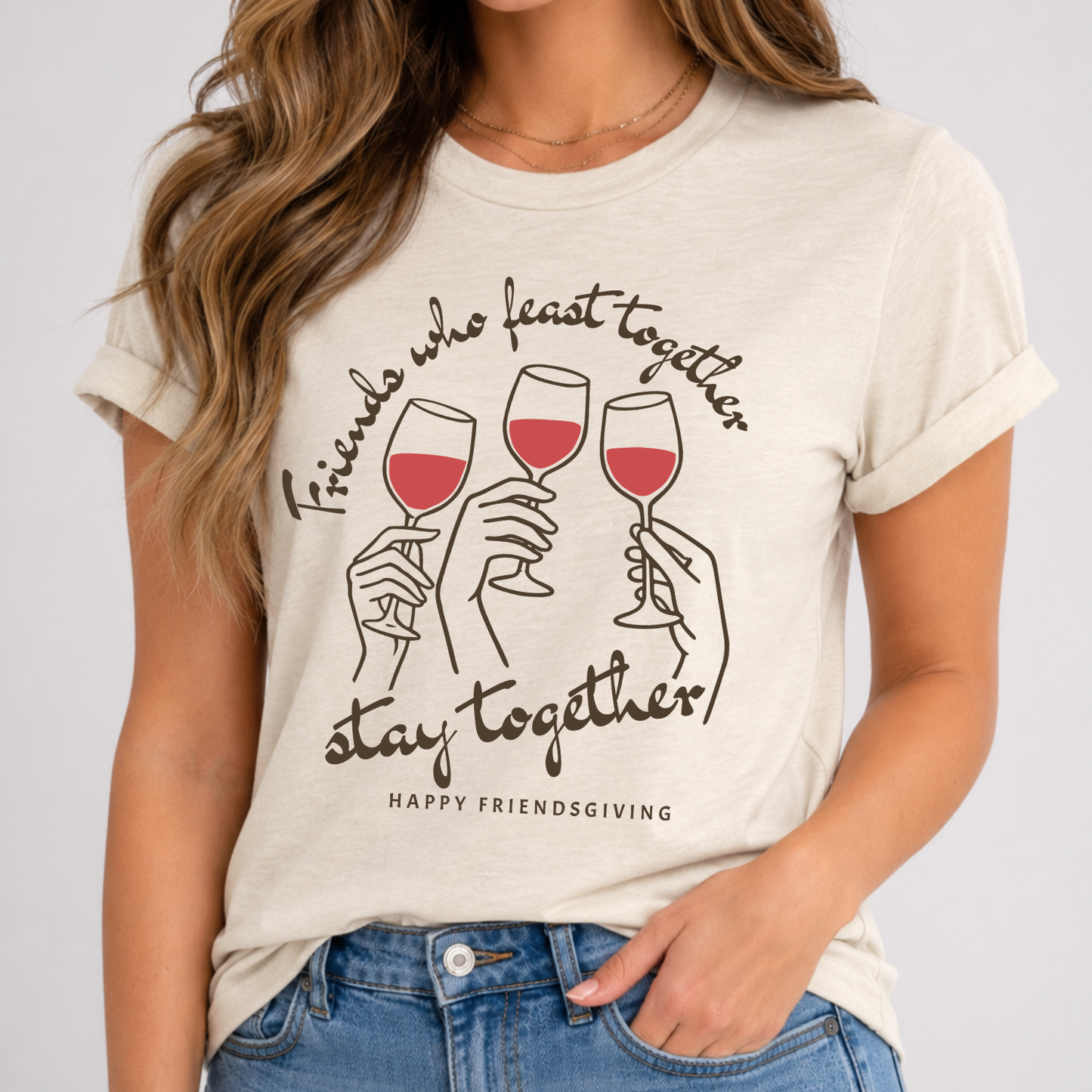 A woman wearing a light beige graphic t-shirt with a 'Friends who feast together stay together Happy Friendsgiving' print featuring three wine glasses, paired with blue jeans.