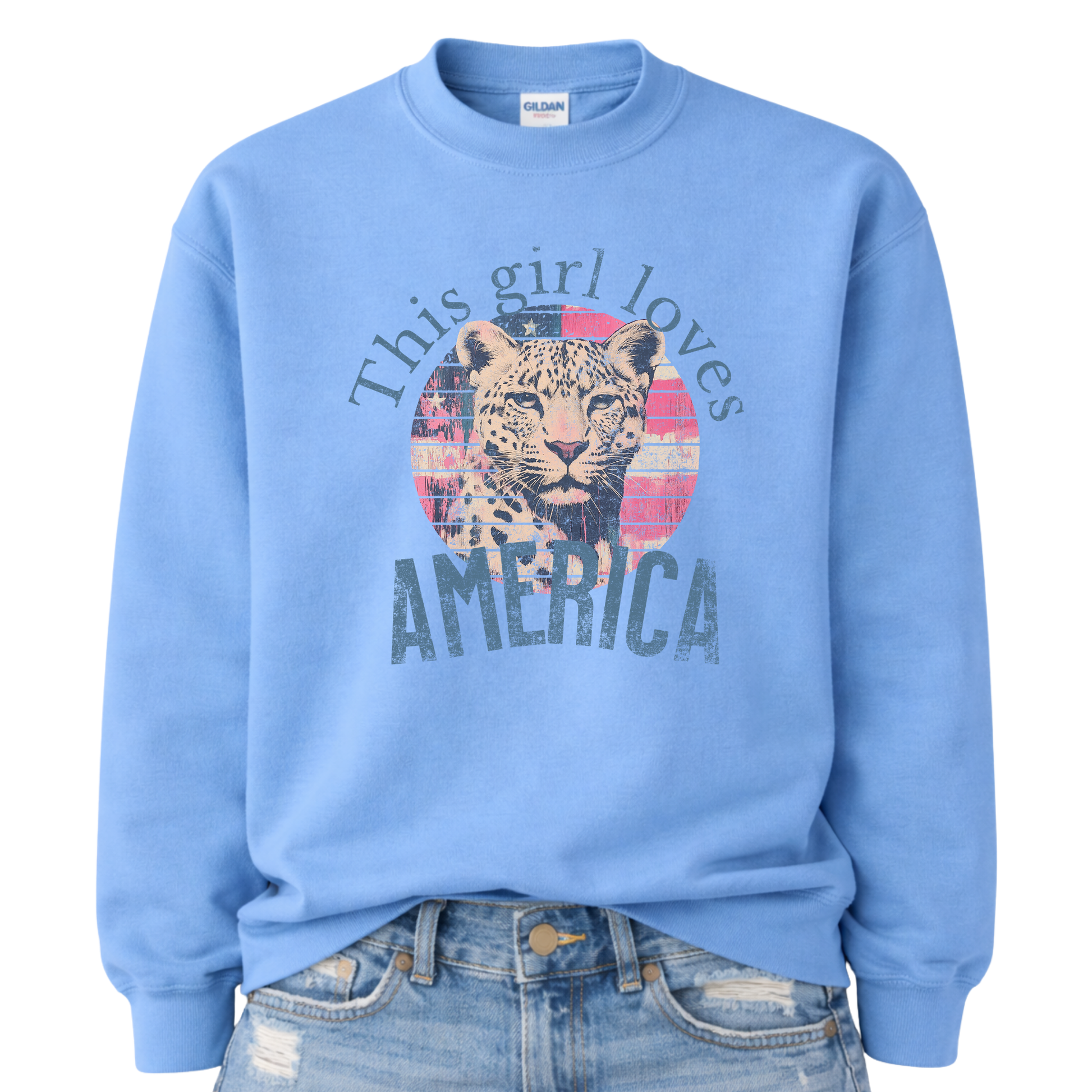 Patriotic leopard graphic on a blue sweatshirt. The design features an illustrated leopard portrait centered over a circular American flag backdrop with distressed red, white, and blue textures. The phrase “This Girl Loves America” curves above and below the artwork in bold, vintage-style lettering. The overall look blends wildlife imagery with Americana elements, creating a strong, retro-inspired patriotic statement. i