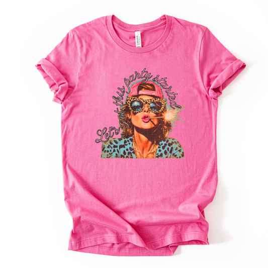 pink Patriotic Fourth of July graphic on a white  t-shirt. The design features an illustrated woman wearing leopard-print sunglasses and a backward cap, holding a lit sparkler with fireworks reflected in the lenses. Curved script lettering adds a playful party vibe, blending bold Americana colors with a modern, fun-forward patriotic style ideal for summer celebrations.