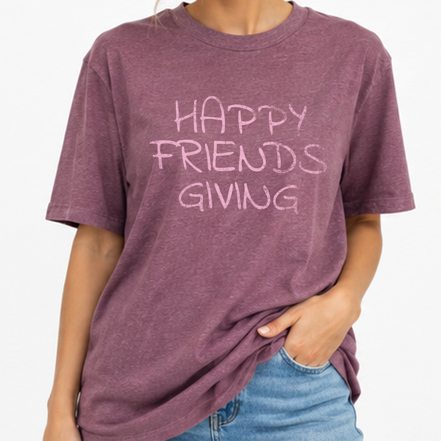 A person wearing a heathered mauve short-sleeve t-shirt with the text 'HAPPY FRIENDS GIVING' printed in a light pink handwritten font across the chest, paired with blue jeans against a plain white background.