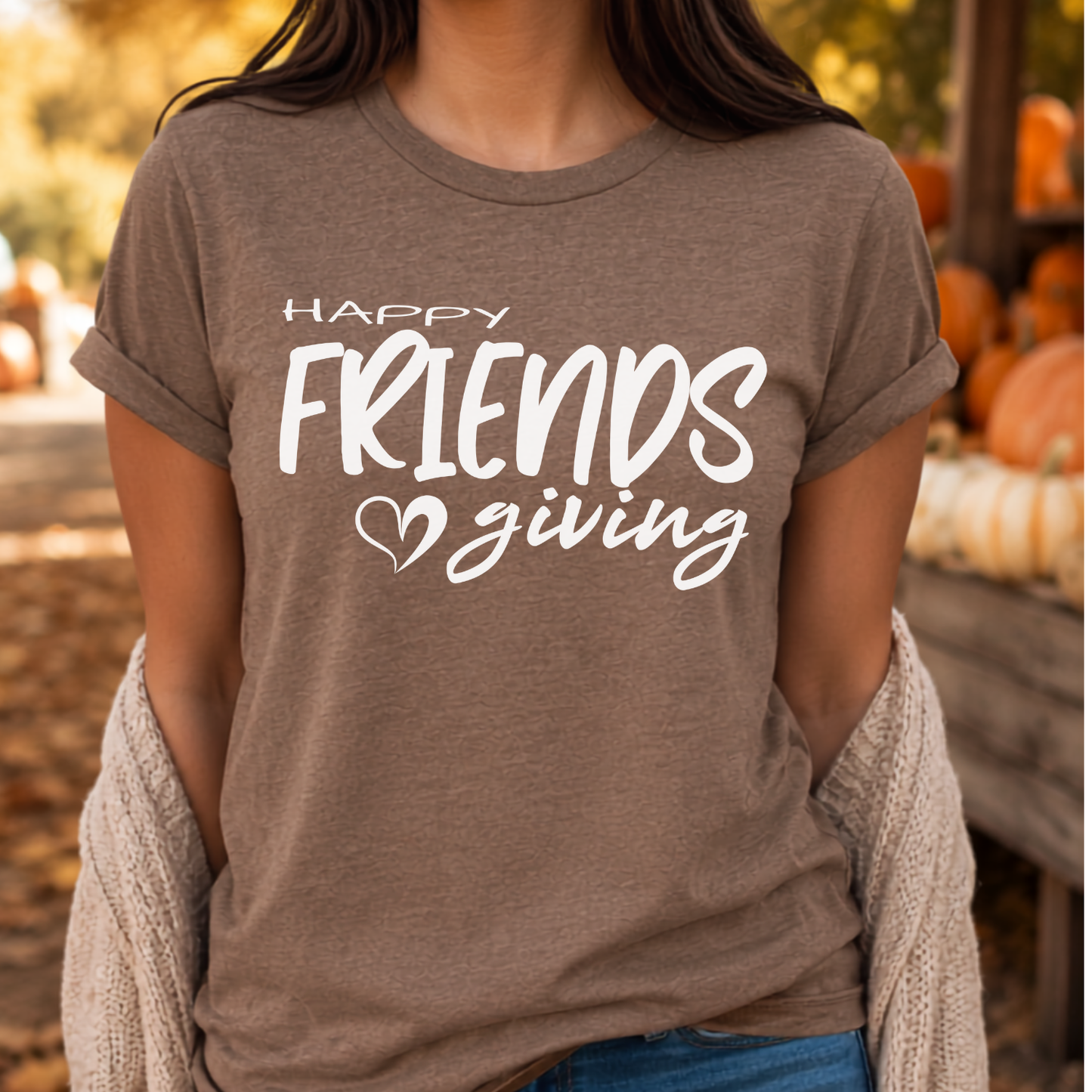 A woman wearing a heather taupe brown graphic t-shirt with the white text 'HAPPY FRIENDS giving' and a small heart, styled with a beige knit cardigan in an autumn-themed setting.