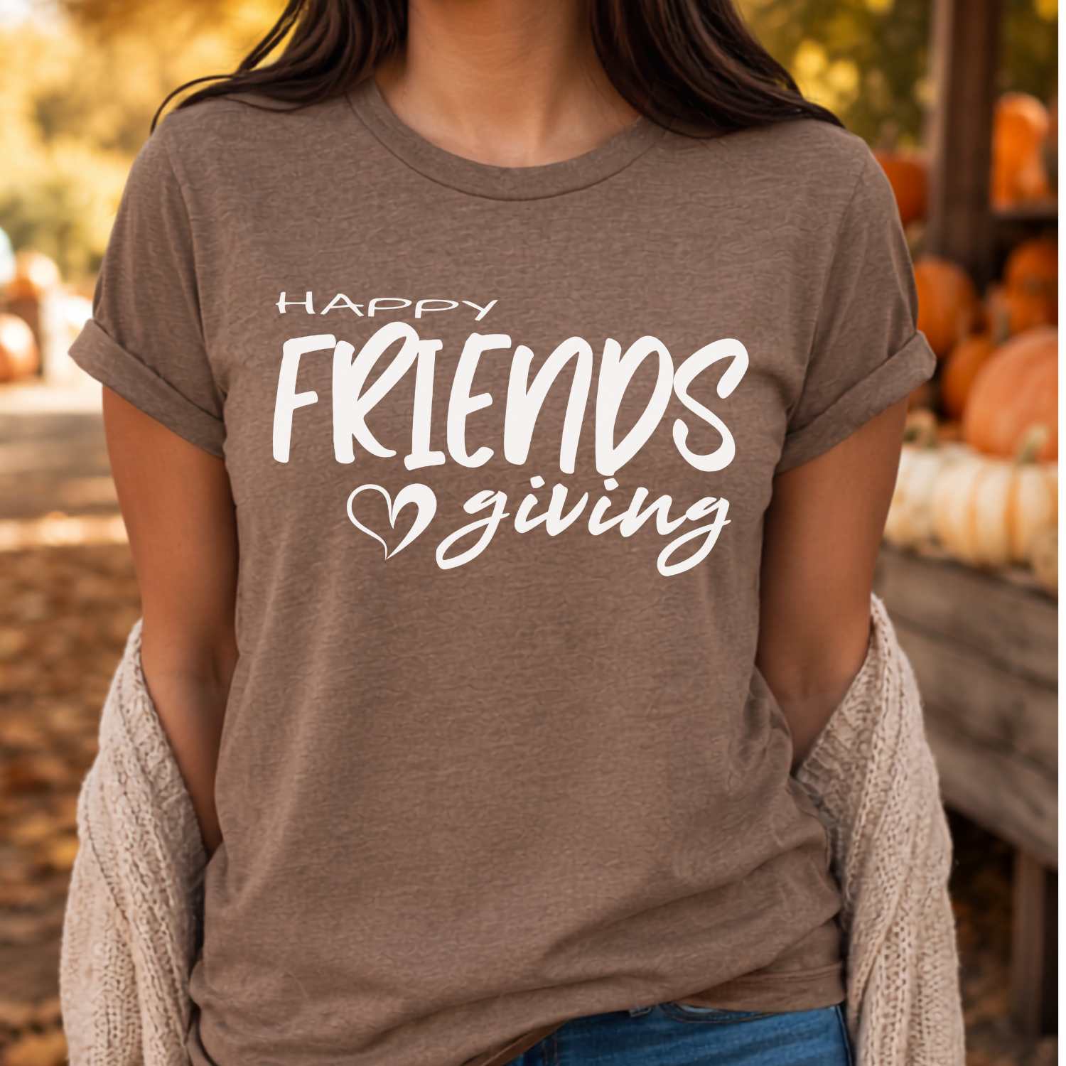 A woman wearing a heather taupe brown graphic t-shirt with the white text 'HAPPY FRIENDS giving' and a small heart, styled with a beige knit cardigan in an autumn-themed setting.