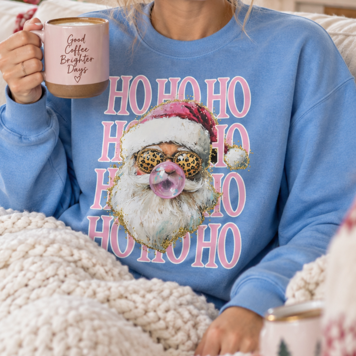 A person wearing a light blue crewneck sweatshirt with a festive graphic of a Santa Claus face wearing leopard-print sunglasses and blowing a pink bubblegum bubble, surrounded by pink 'HO HO HO' text. The person is holding a pink ceramic mug with 'Good Coffee Brighter Days' written on it, wrapped in a chunky cream knit blanket.