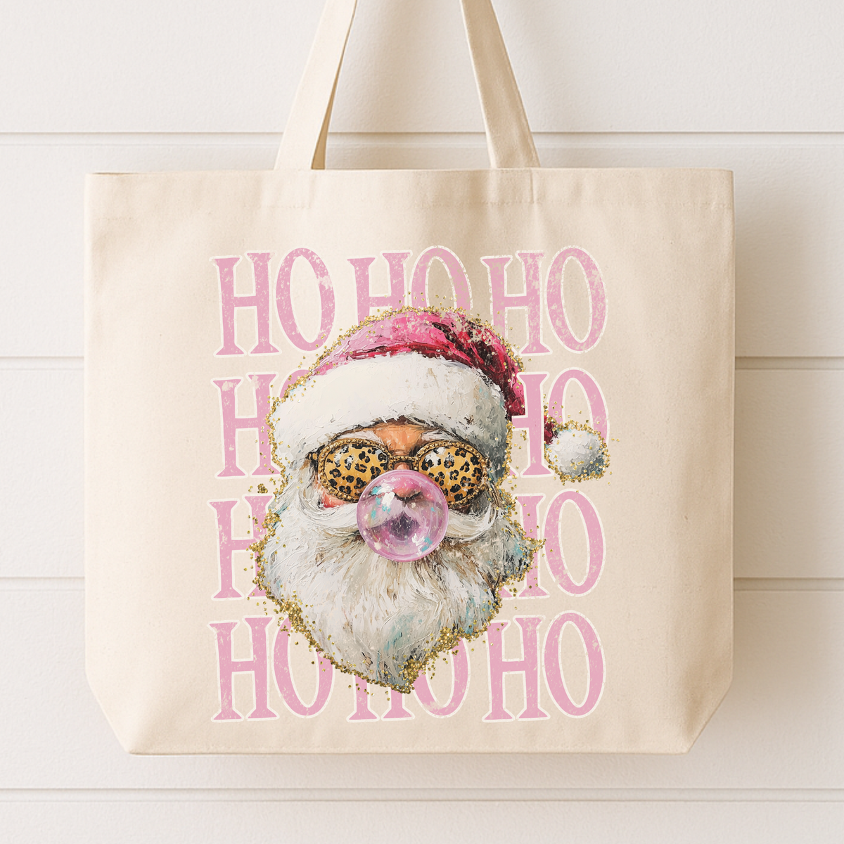 A cream canvas tote bag with a festive graphic print of a Santa Claus face wearing leopard-print sunglasses and blowing a bubblegum bubble, set against a background of pink 'HO HO HO' text.