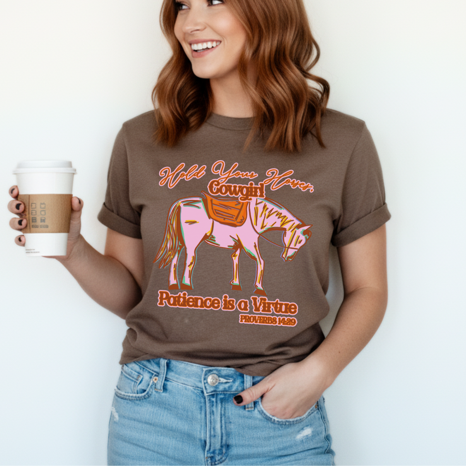 Woman wearing a  Bella Canvas graphic t-shirt featuring a colorful cowgirl horse illustration with the phrase “Hold Your Horses, Cowgirl” and “Patience is a Virtue Proverbs 14:29,” styled casually with jeans and coffee cup for a relaxed, western-inspired look.