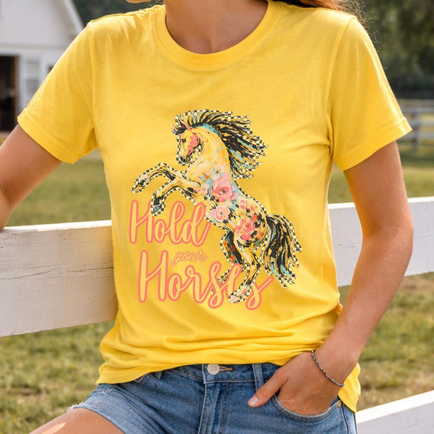 Person wearing a yellow t-shirt with a horse graphic and text, sitting on a wooden fence.  Shirt says Hold your horses