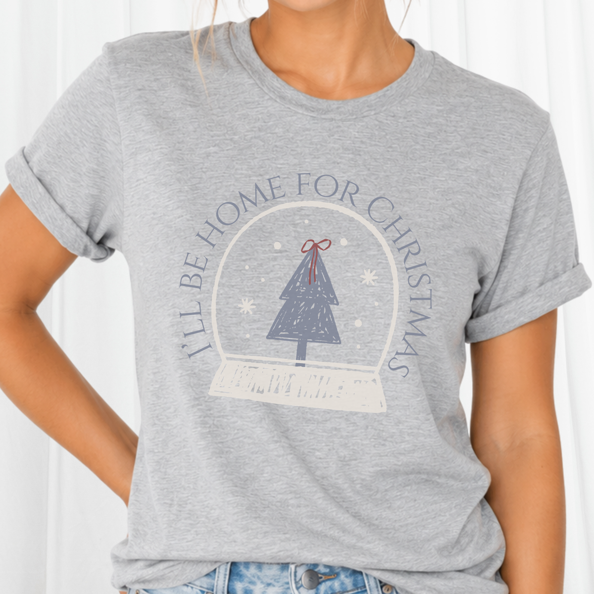 A sweet holiday graphic featuring a doodle style art snow globe, Christmas tree, and the saying “I’ll Be Home for Christmas.” on a gray tee