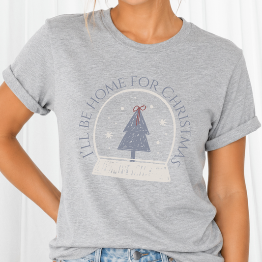 A sweet holiday graphic featuring a doodle style art snow globe, Christmas tree, and the saying “I’ll Be Home for Christmas.” on a gray tee