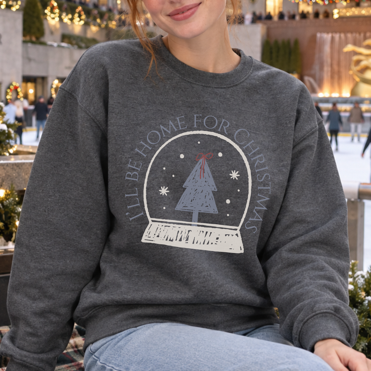 A woman wearing a dark gray crewneck sweatshirt with a graphic of a Christmas tree inside a snow globe and the text 'I'LL BE HOME FOR CHRISTMAS' in light gray, set against a blurred festive outdoor background.