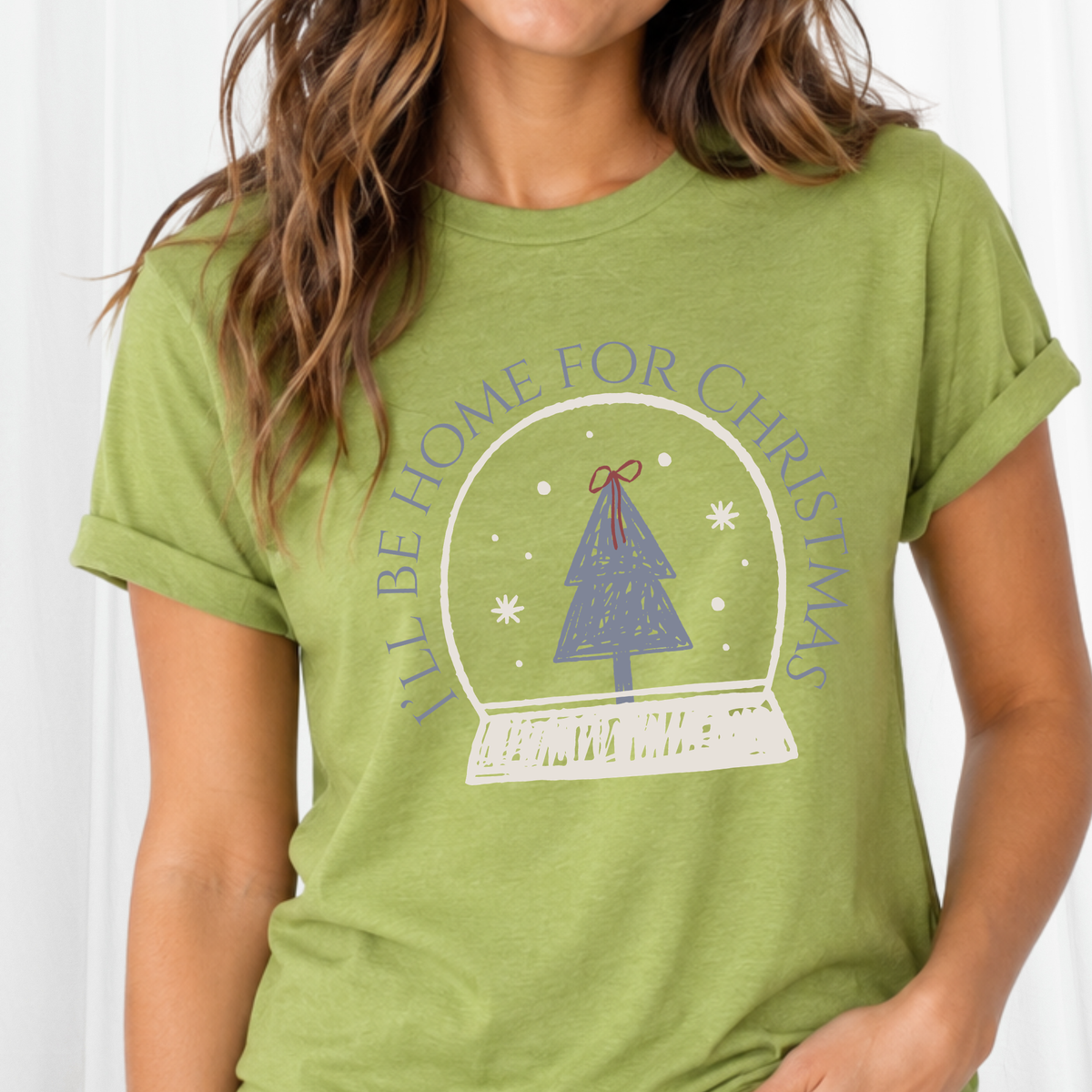A sweet holiday graphic featuring a doodle style art snow globe, Christmas tree, and the saying “I’ll Be Home for Christmas.” on a heather green tee