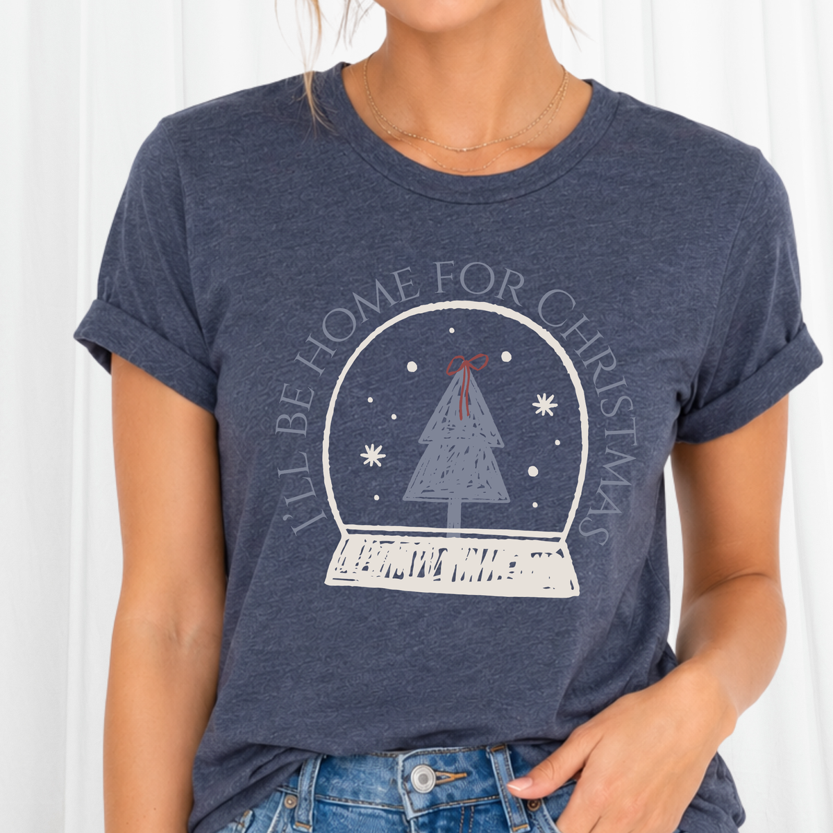 A sweet holiday graphic featuring a doodle style art snow globe, Christmas tree, and the saying “I’ll Be Home for Christmas.” on a heather navy tee