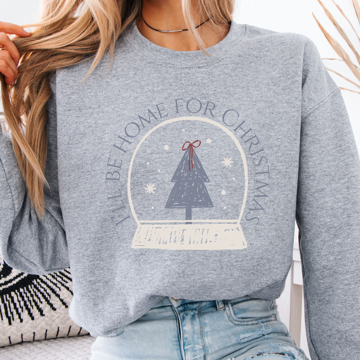 A sweet holiday graphic featuring a hand-drawn snow globe, Christmas tree, and the saying “I’ll Be Home for Christmas.” on ash gray sweatshirt