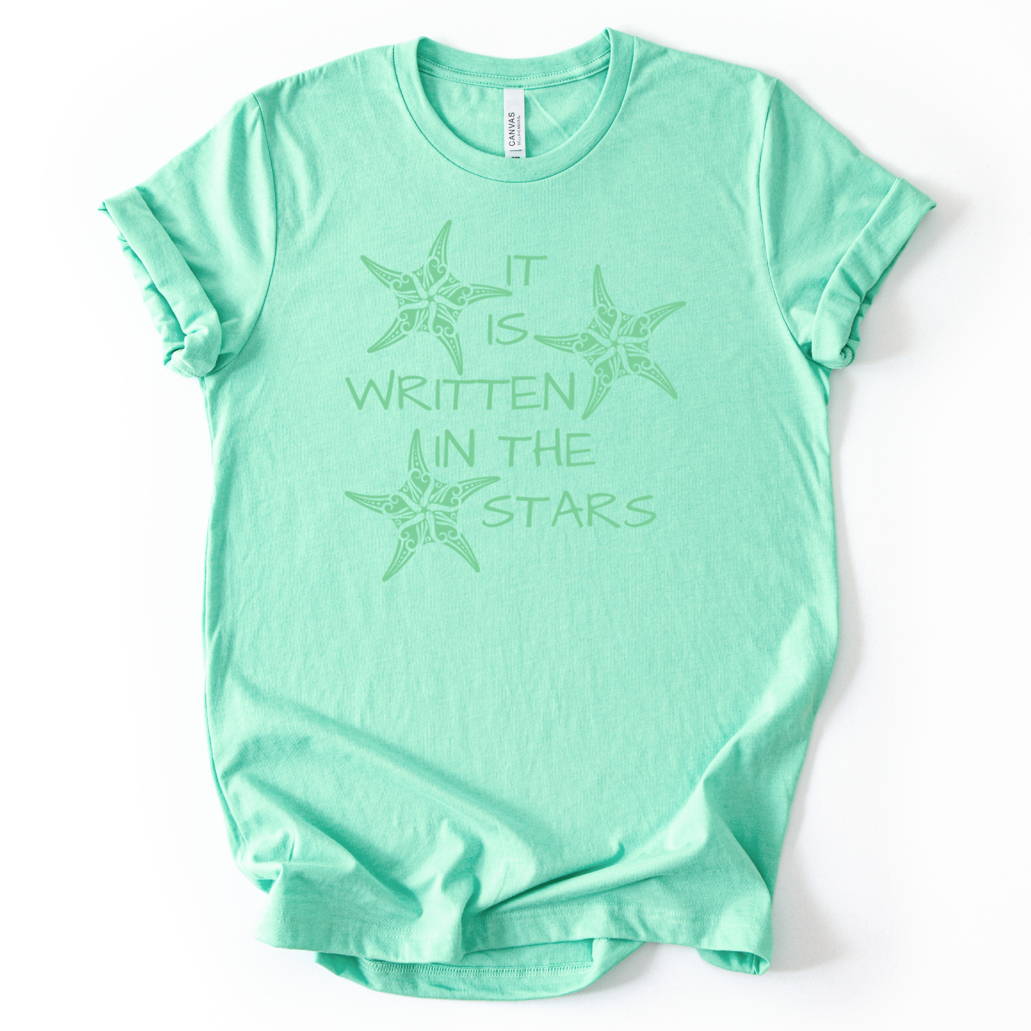 Coastal beach graphic on a soft unisex heather mint graphic tee. The design features the phrase It Is Written In The Stars surrounded by illustrated sea stars. The starfish artwork includes decorative coastal pattern details inside the shapes, giving the illustration a textured ocean inspired look. 