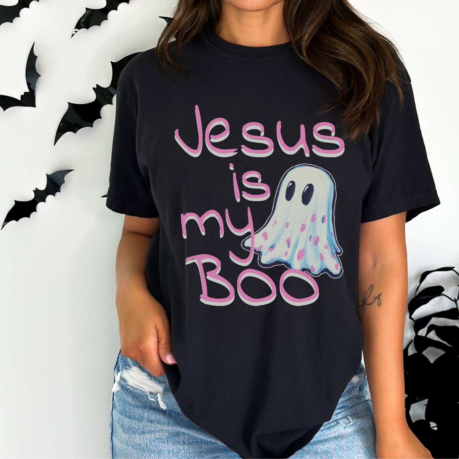 A woman wearing a black graphic t-shirt with the text 'Jesus is my Boo' in pink and a ghost graphic, paired with light-wash distressed jeans. The background is white with black bat decorations.