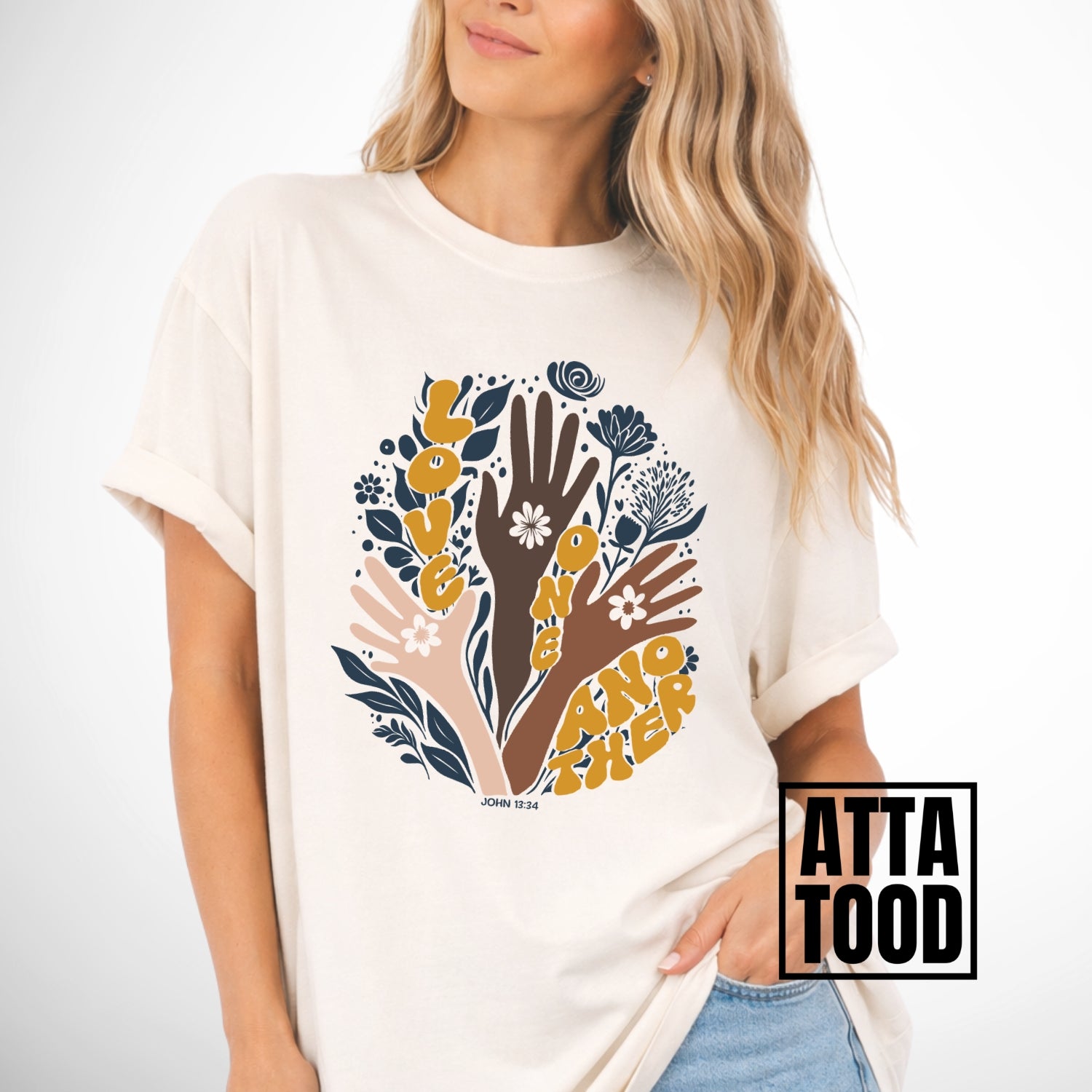 Person wearing a ivory t-shirt with a colorful graphic of hands and flowers on a white background. John 13 Love One Another Comfort Colors Graphic Tee 