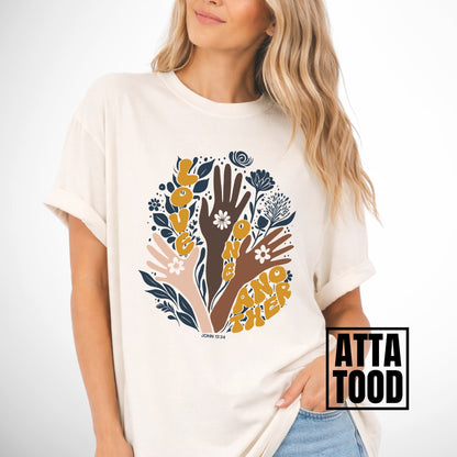 Person wearing a ivory t-shirt with a colorful graphic of hands and flowers on a white background. John 13 Love One Another Comfort Colors Graphic Tee 