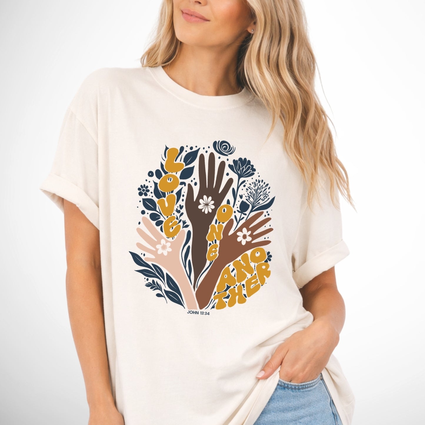 Woman wearing a ivory t-shirt with a graphic design of hands and flowers on a plain background. John 13 Love One Another Comfort Colors Graphic Tee 