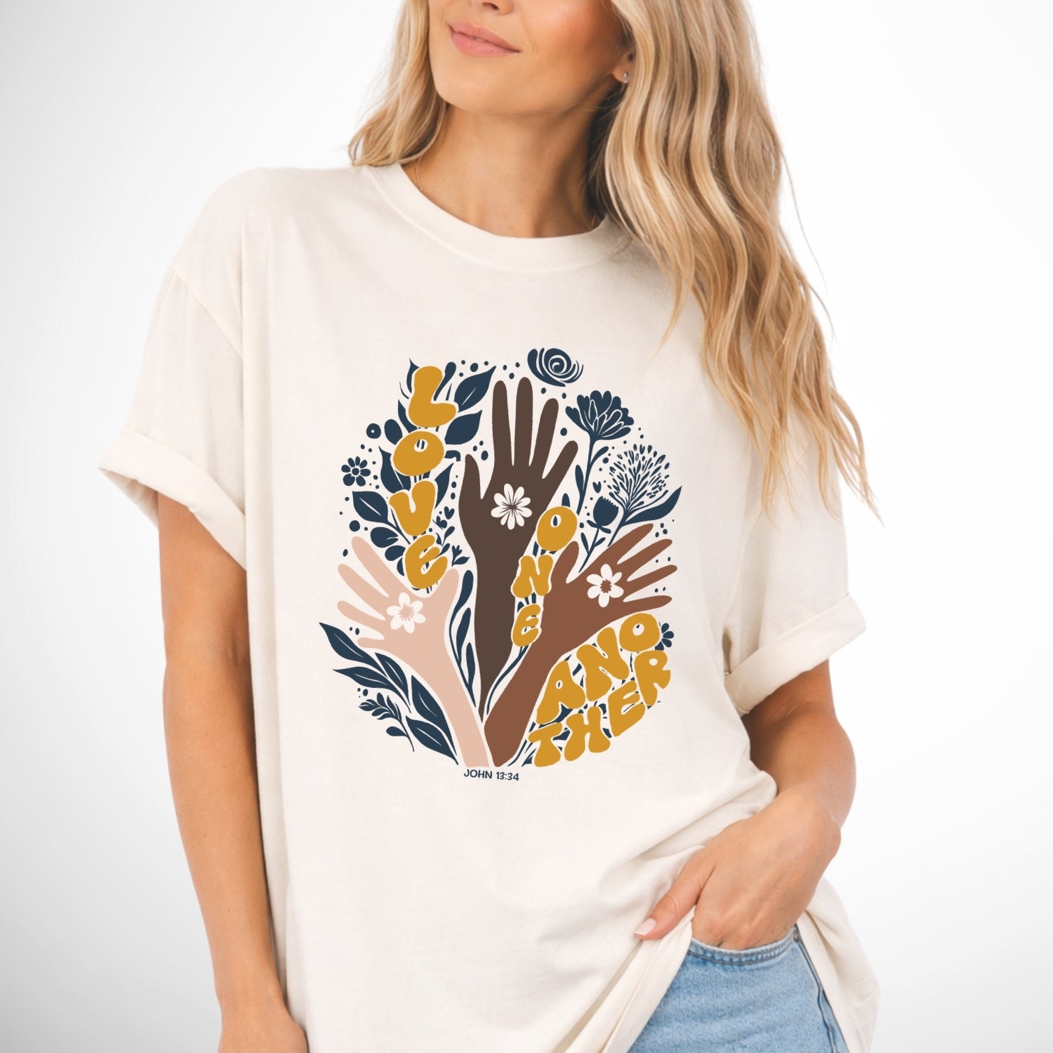 Woman wearing a ivory t-shirt with a graphic design of hands and flowers on a plain background. John 13 Love One Another Comfort Colors Graphic Tee 
