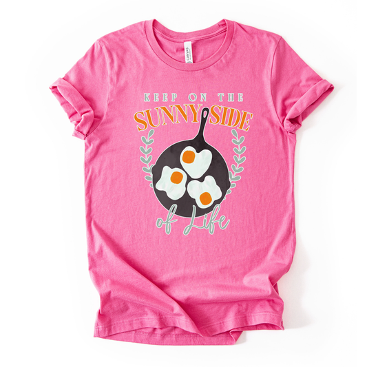 Bella Canvas unisex graphic t-shirt mockup with “Keep On The Sunny Side of Life” text and a sunny side up eggs design in a black skillet, with small leafy accents; bright, cheerful, retro-style positive quote tee.