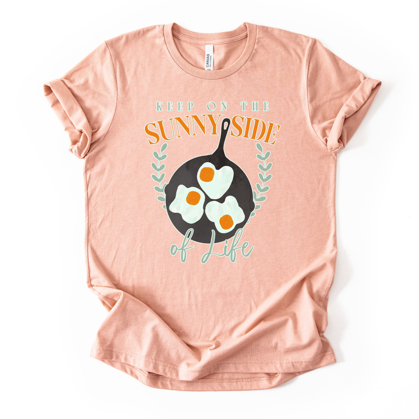 Bella Canvas unisex graphic t-shirt mockup with “Keep On The Sunny Side of Life” text and a sunny side up eggs design in a black skillet, with small leafy accents; bright, cheerful, retro-style positive quote tee.