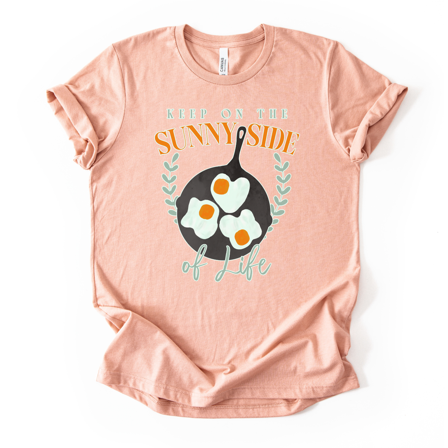 Bella Canvas unisex graphic t-shirt mockup with “Keep On The Sunny Side of Life” text and a sunny side up eggs design in a black skillet, with small leafy accents; bright, cheerful, retro-style positive quote tee.