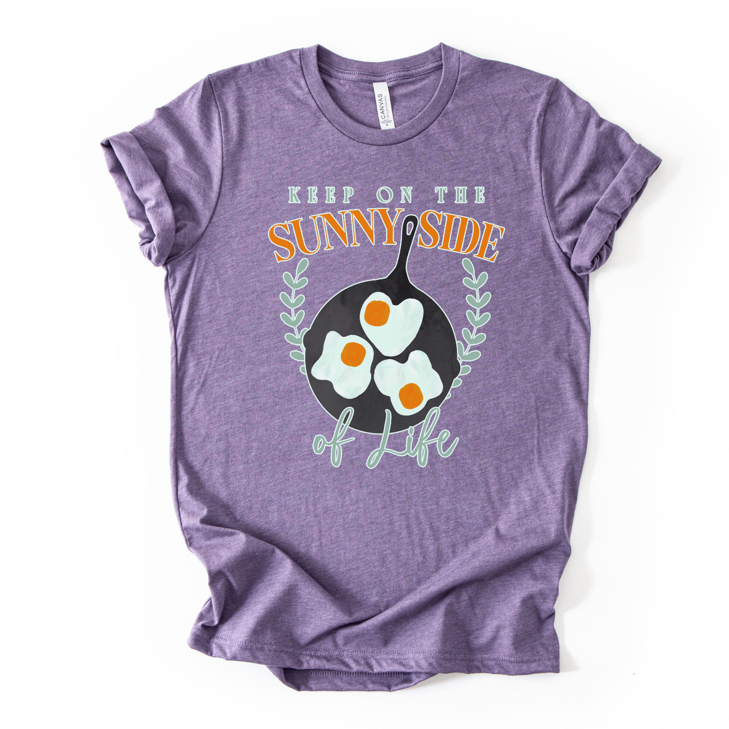 Bella Canvas unisex graphic t-shirt mockup with “Keep On The Sunny Side of Life” text and a sunny side up eggs design in a black skillet, with small leafy accents; bright, cheerful, retro-style positive quote tee.