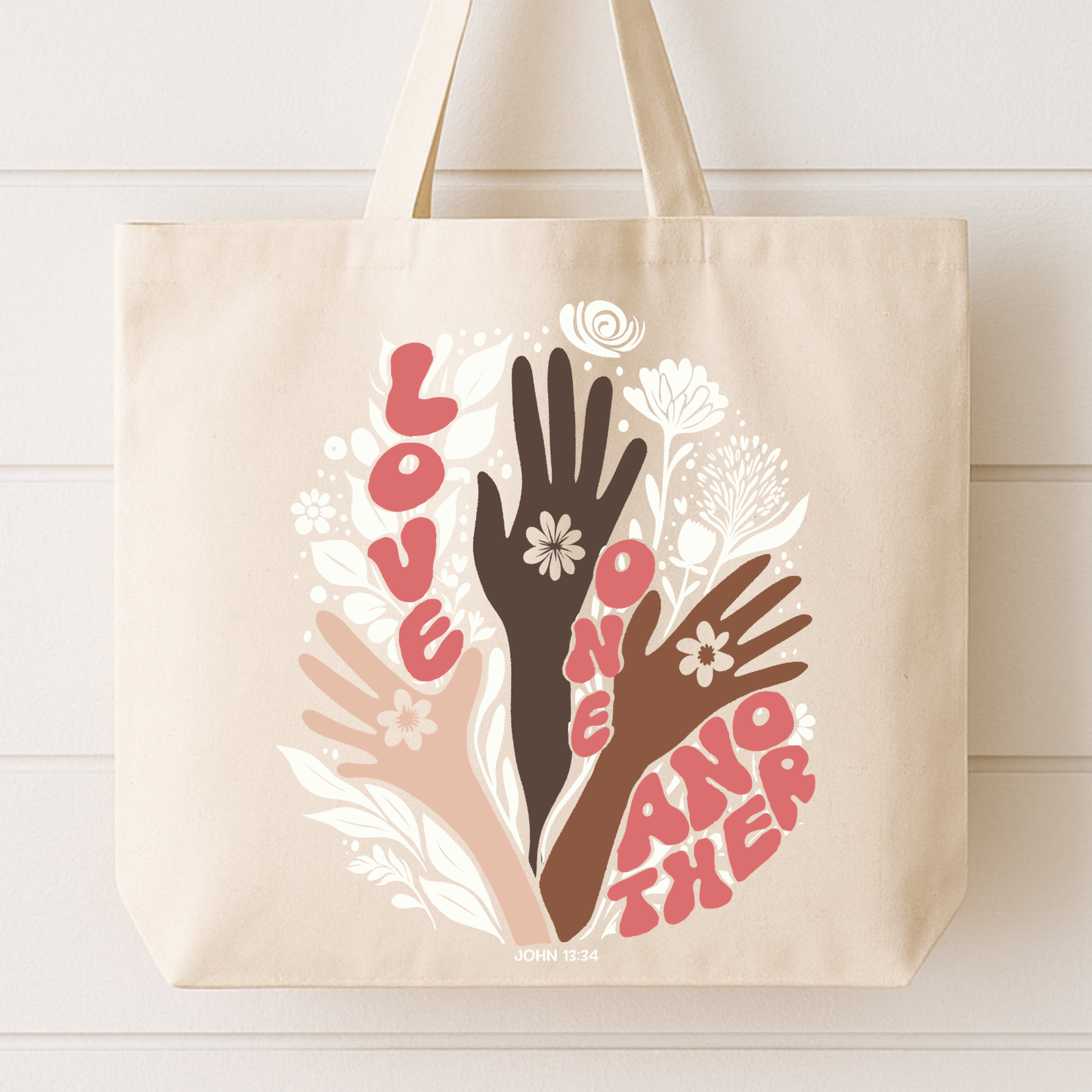 Beige tote bag with artistic depiction of hands and flowers, featuring the text 'Love One Another'.