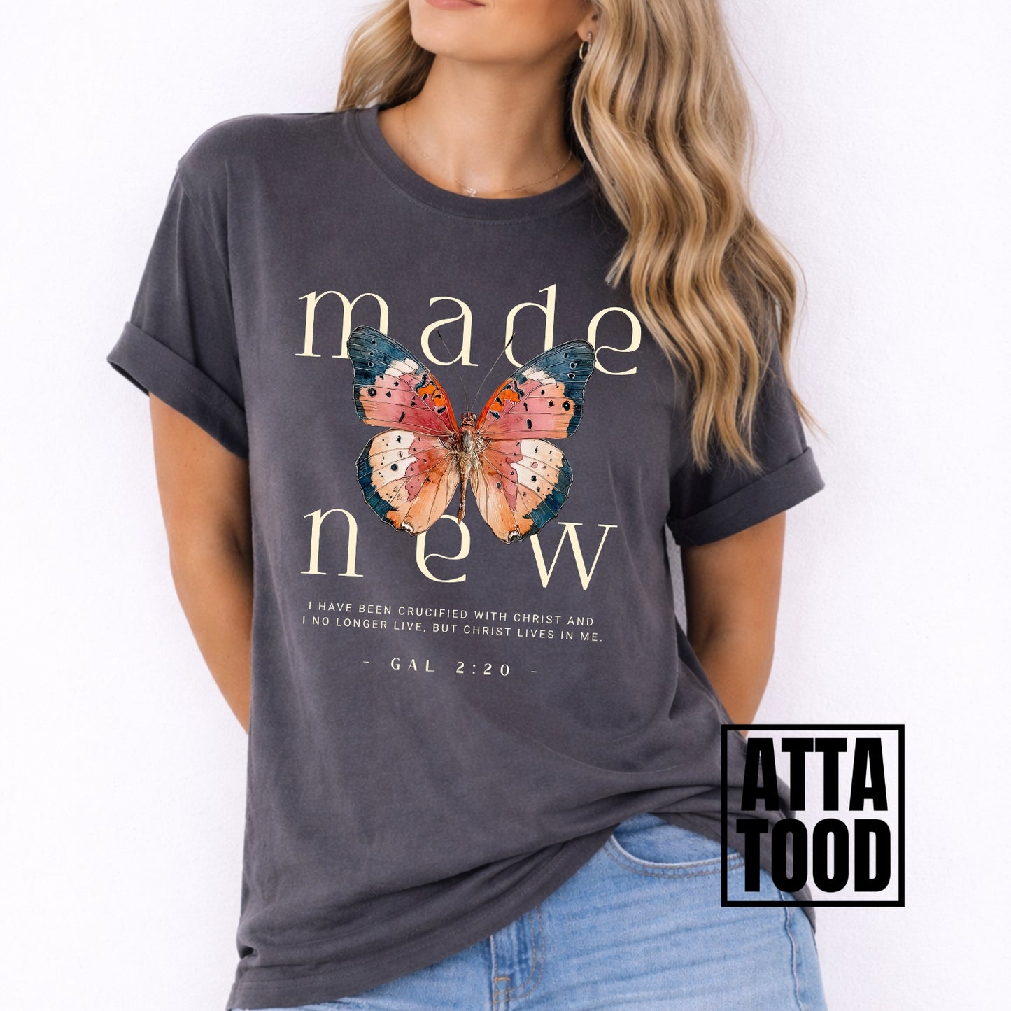 Woman wearing a dark gray t-shirt with a butterfly graphic and text, brand 'Atta Tood' visible. Made new Galatians 2:20