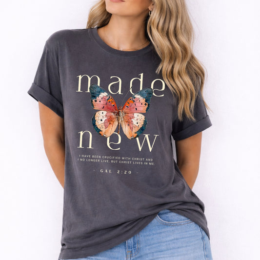 Person wearing a dark gray t-shirt with butterfly design and text on a white background. Made New Bible Verse Comfort Colors Butterfly Graphic Tee
