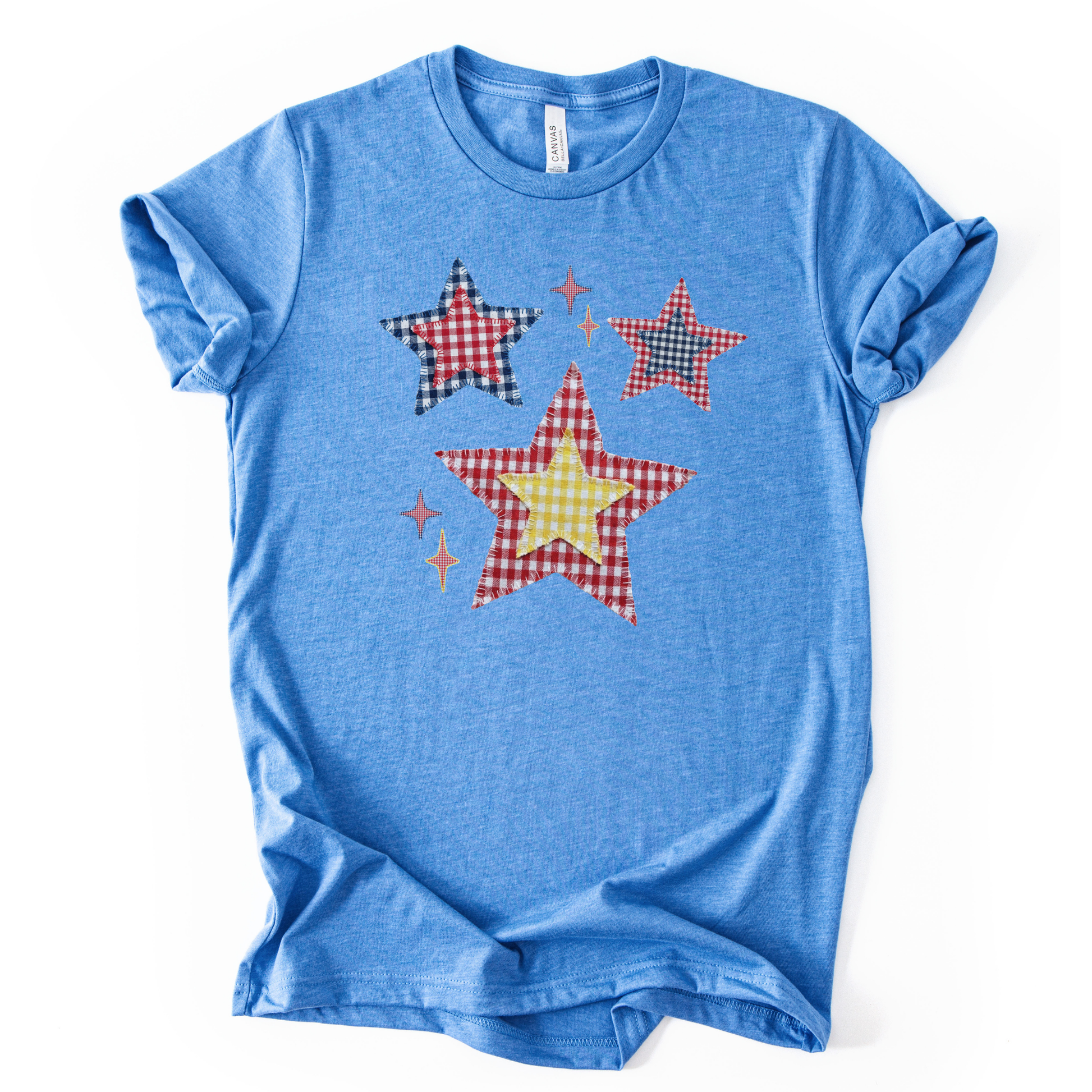 Patriotic Americana star graphic on a blue t-shirt. The design features illustrated patchwork stars arranged in a playful cluster, using red, white, and blue gingham-style patterns with subtle texture and stitching detail. Small accent stars add movement and balance around the main shapes. The overall look feels classic, cheerful, and vintage-inspired, making it ideal for Fourth of July, summer Americana collections, and everyday patriotic wear.