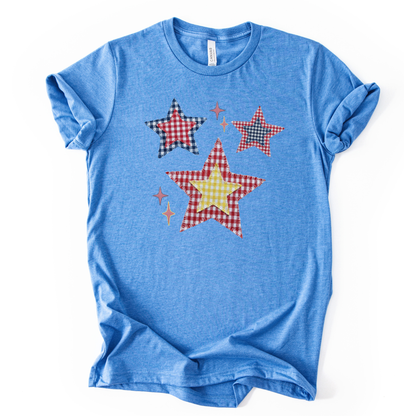 Patriotic Americana star graphic on a blue t-shirt. The design features illustrated patchwork stars arranged in a playful cluster, using red, white, and blue gingham-style patterns with subtle texture and stitching detail. Small accent stars add movement and balance around the main shapes. The overall look feels classic, cheerful, and vintage-inspired, making it ideal for Fourth of July, summer Americana collections, and everyday patriotic wear.