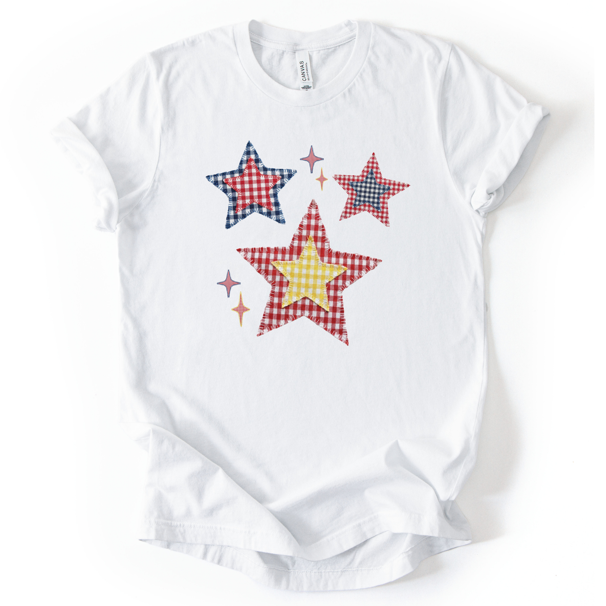 Patriotic Americana star graphic on a white t-shirt. The design features illustrated patchwork stars arranged in a playful cluster, using red, white, and blue gingham-style patterns with subtle texture and stitching detail. Small accent stars add movement and balance around the main shapes. The overall look feels classic, cheerful, and vintage-inspired, making it ideal for Fourth of July, summer Americana collections, and everyday patriotic wear.