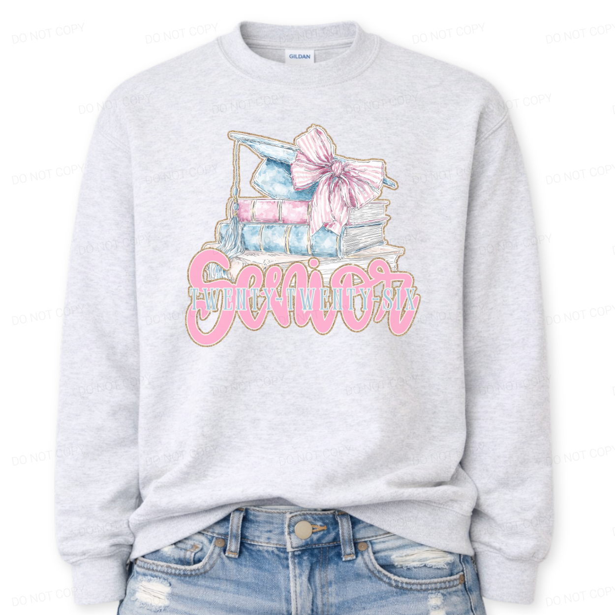 ash Graduation 2026 graphic on a classic crewneck sweatshirt. The design features a stacked book illustration in soft blue and blush tones topped with a graduation cap and oversized pink bow. Bold Senior Twenty Twenty Six lettering anchors the artwork, giving it a strong Class of 2026 presence with a preppy feminine academic aesthetic that works for both high school seniors and college graduates.