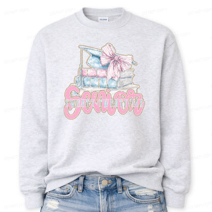 ash Graduation 2026 graphic on a classic crewneck sweatshirt. The design features a stacked book illustration in soft blue and blush tones topped with a graduation cap and oversized pink bow. Bold Senior Twenty Twenty Six lettering anchors the artwork, giving it a strong Class of 2026 presence with a preppy feminine academic aesthetic that works for both high school seniors and college graduates.