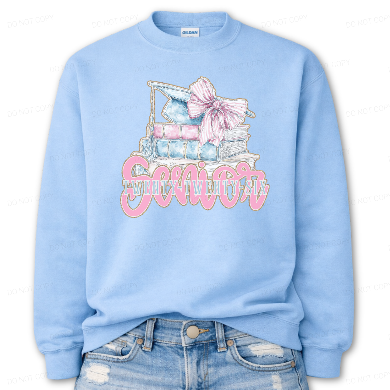 Graduation 2026 graphic on a classic crewneck sweatshirt. The design features a stacked book illustration in soft blue and blush tones topped with a graduation cap and oversized pink bow. Bold Senior Twenty Twenty Six lettering anchors the artwork, giving it a strong Class of 2026 presence with a preppy feminine academic aesthetic that works for both high school seniors and college graduates.