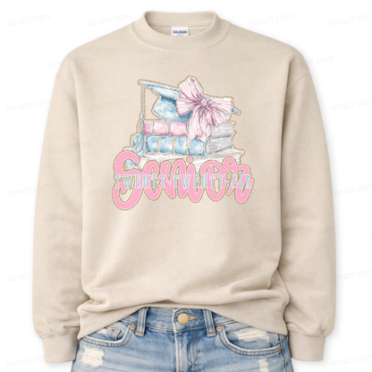 Graduation 2026 graphic on a classic sand crewneck sweatshirt. The design features a stacked book illustration in soft blue and blush tones topped with a graduation cap and oversized pink bow. Bold Senior Twenty Twenty Six lettering anchors the artwork, giving it a strong Class of 2026 presence with a preppy feminine academic aesthetic that works for both high school seniors and college graduates.