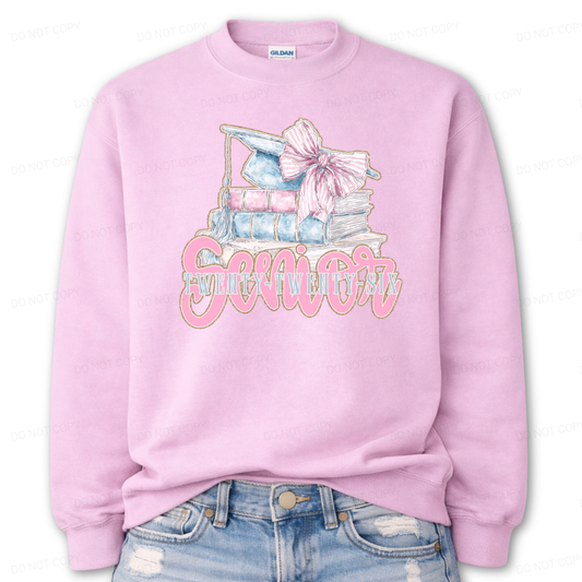 Graduation 2026 graphic on a classic pink crewneck sweatshirt. The design features a stacked book illustration in soft blue and blush tones topped with a graduation cap and oversized pink bow. Bold Senior Twenty Twenty Six lettering anchors the artwork, giving it a strong Class of 2026 presence with a preppy feminine academic aesthetic that works for both high school seniors and college graduates.