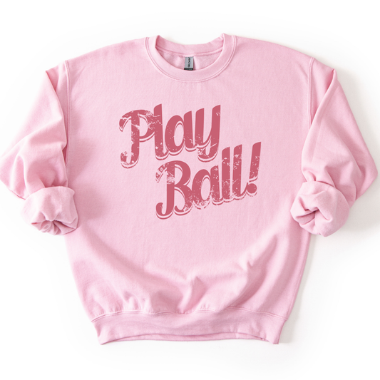 Light pink crewneck sweatshirt featuring distressed retro script text reading “Play Ball,” styled flat with sleeves scrunched