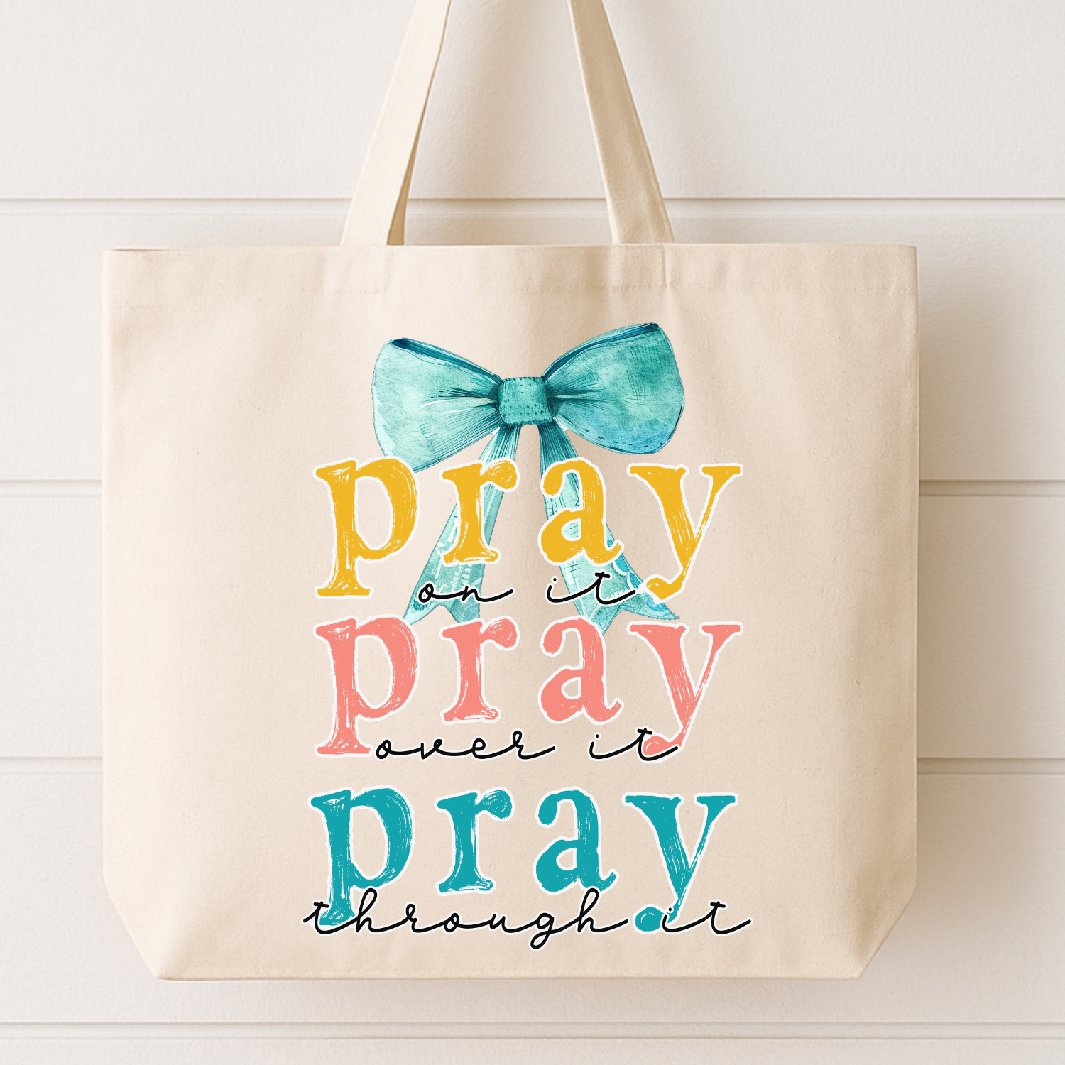 Beige tote bag with colorful text and a bow design on a white wooden surface. Pray on it pray over it pray through it