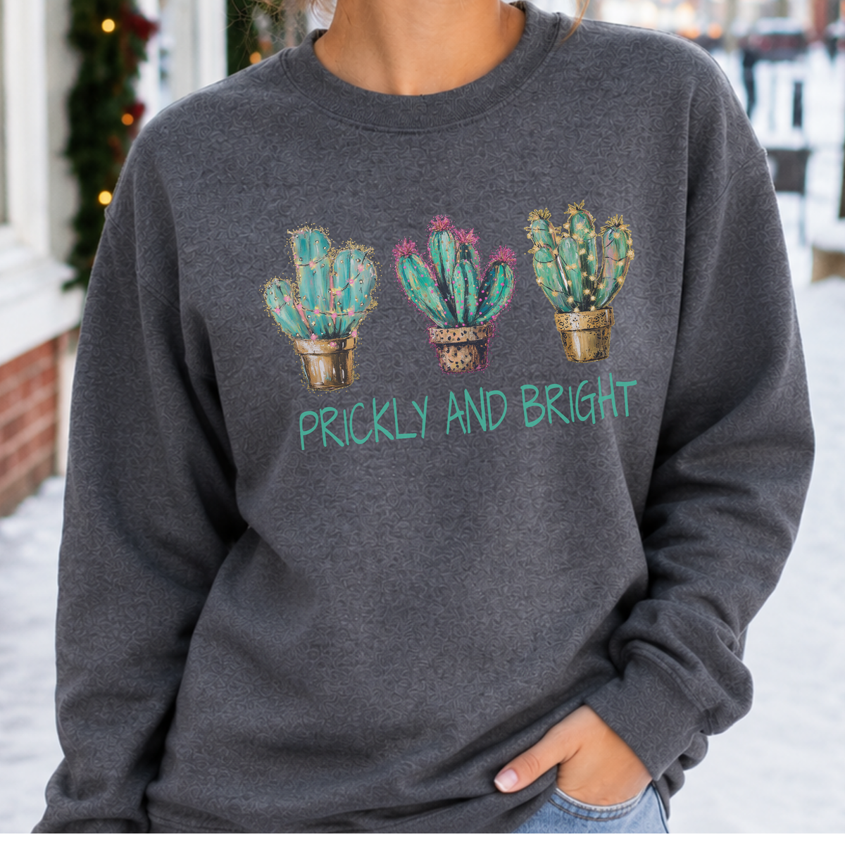 A person wearing a dark heather gray crewneck sweatshirt with a graphic of three potted cacti and the text 'PRICKLY AND BRIGHT' in teal, photographed outdoors in a snowy setting.