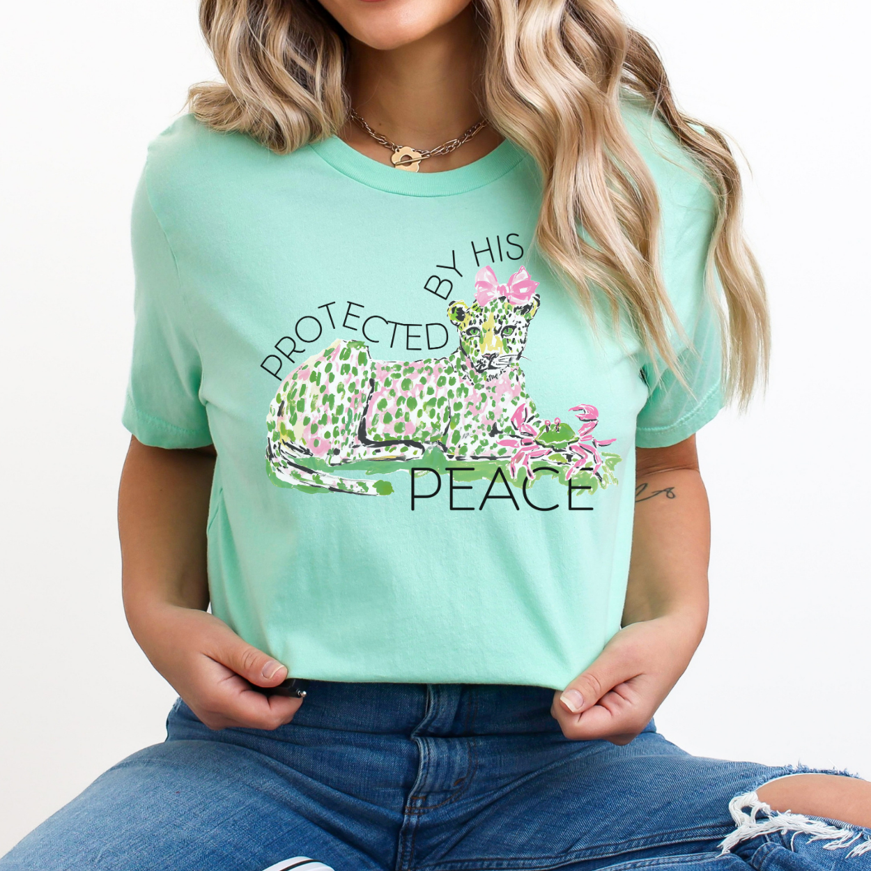 Christian faith graphic on a Bella Canvas t-shirt. The design features an illustrated leopard resting peacefully with soft green and pink pattern details, accented with a pink bow and floral elements. The phrase “Protected by His Peace” arches above and below the artwork in clean lettering, blending feminine spring style with a calm, faith-forward message.