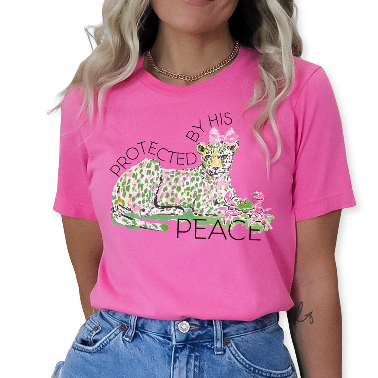 Christian faith graphic on a Bella Canvas t-shirt. The design features an illustrated leopard resting peacefully with soft green and pink pattern details, accented with a pink bow and floral elements. The phrase “Protected by His Peace” arches above and below the artwork in clean lettering, blending feminine spring style with a calm, faith-forward message.