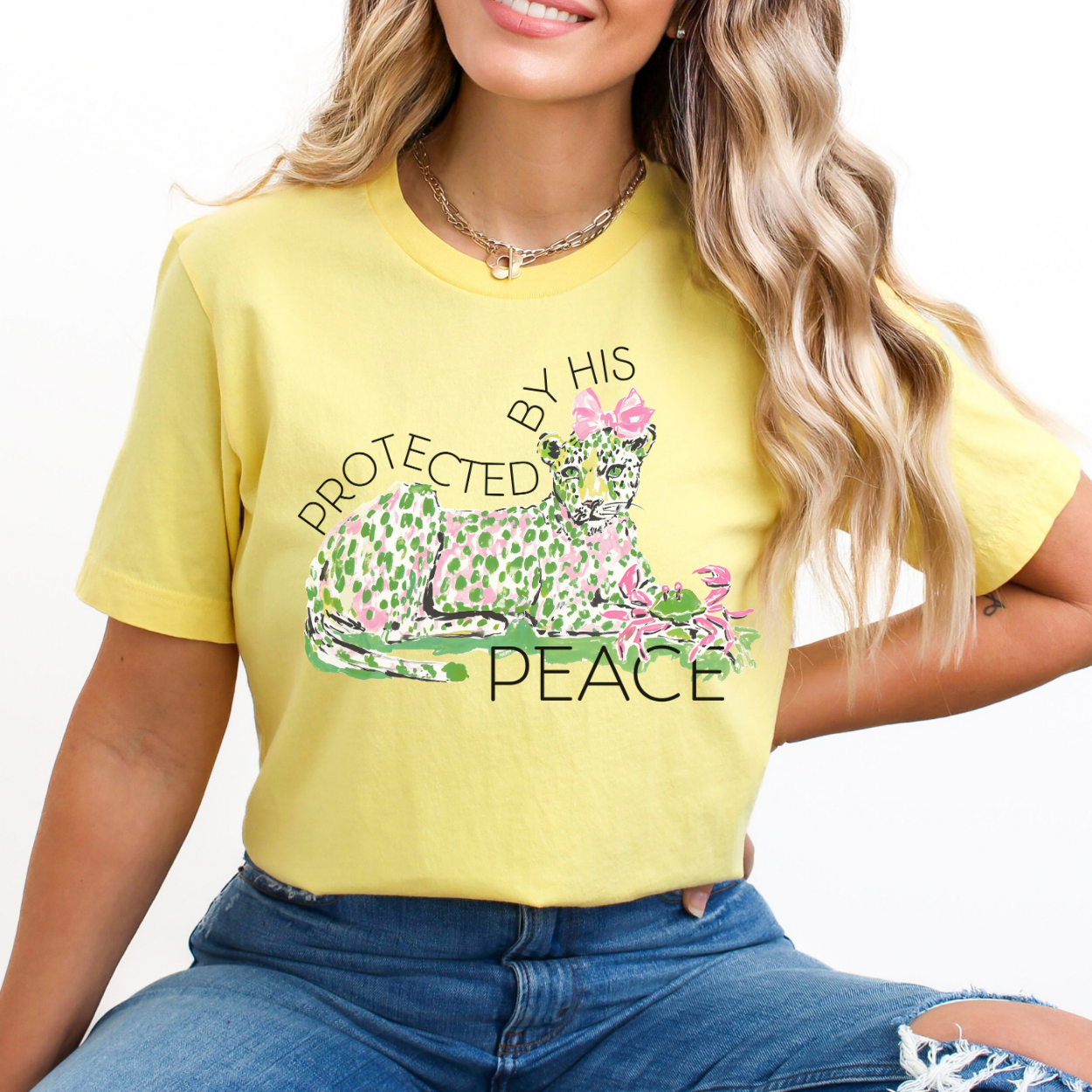 Christian faith graphic on a Bella Canvas t-shirt. The design features an illustrated leopard resting peacefully with soft green and pink pattern details, accented with a pink bow and floral elements. The phrase “Protected by His Peace” arches above and below the artwork in clean lettering, blending feminine spring style with a calm, faith-forward message.