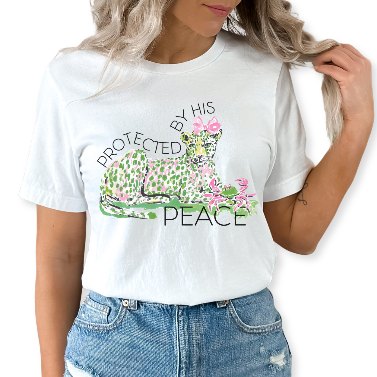 Christian faith graphic on a Bella Canvas t-shirt. The design features an illustrated leopard resting peacefully with soft green and pink pattern details, accented with a pink bow and floral elements. The phrase “Protected by His Peace” arches above and below the artwork in clean lettering, blending feminine spring style with a calm, faith-forward message.