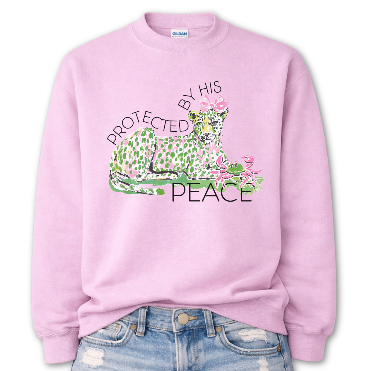 Pink Bold Christian Easter graphic on a Gildan crewneck sweatshirt. The design features a reclining leopard illustrated in soft pink and green tones with a coordinating bow, paired with the phrase “Protected By His Peace” in a clean, modern layout. The artwork blends feminine spring colors with a strong faith message