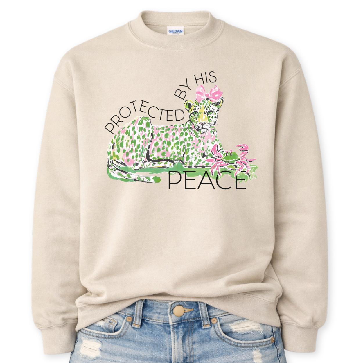 Beige Bold Christian Easter graphic on a Gildan crewneck sweatshirt. The design features a reclining leopard illustrated in soft pink and green tones with a coordinating bow, paired with the phrase “Protected By His Peace” in a clean, modern layout. The artwork blends feminine spring colors with a strong faith message