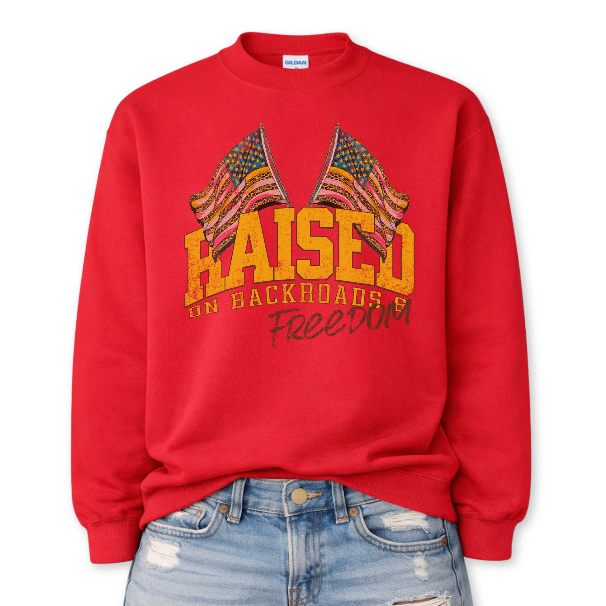 Patriotic freedom graphic on a red sweatshirt. The design features bold block lettering that says "Raised on backroads and freedom" paired with illustrated American flags positioned above the text, finished with a distressed, vintage-inspired texture. The layout blends classic Americana elements with a laid-back, all-American feel, creating a strong patriotic statement