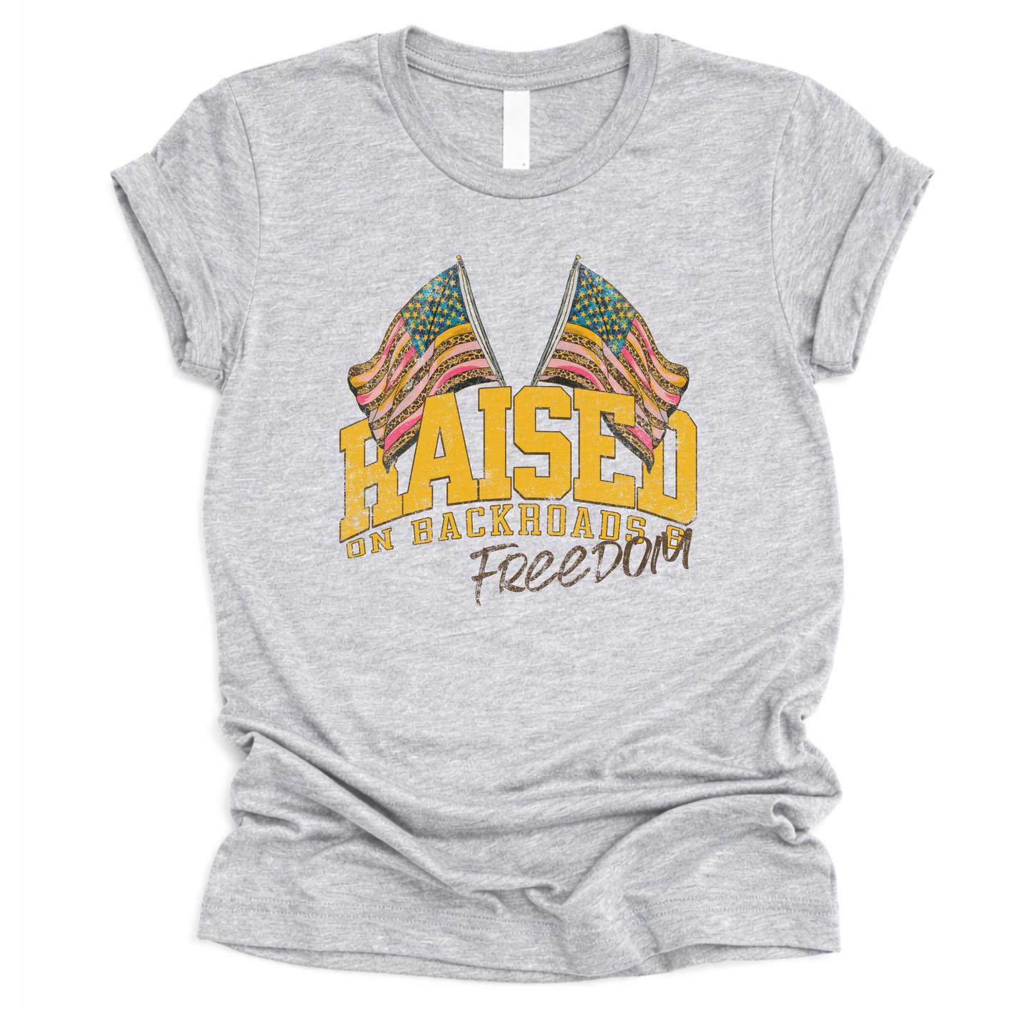 Patriotic Americana graphic on a t-shirt. The design features bold varsity-style “RAISED” lettering with crossed American flags above, finished with “on backroads” and “Freedom” in script below. The distressed, vintage-inspired texture and warm tones make it an easy Fourth of July t-shirt.  gray color tee